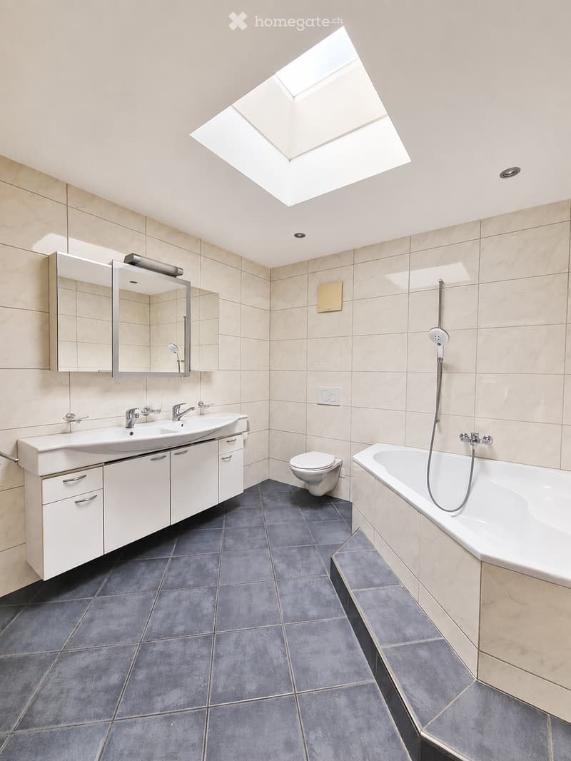 Modern bathroom with double sink, large mirror, bathtub, toilet, and skylight window providing natural light.