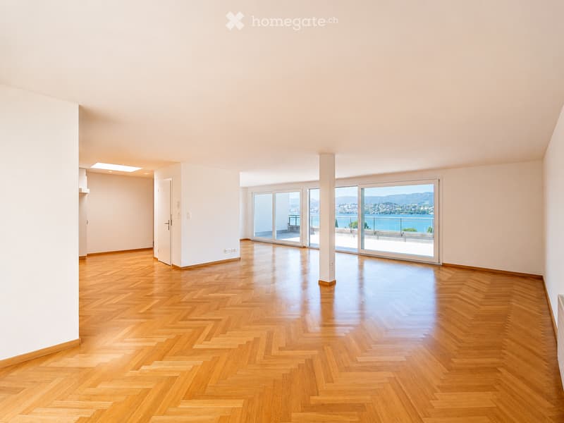 Empty room with wooden herringbone flooring, white walls, large sliding glass doors leading to a balcony with a scenic view of the sea and mountains.