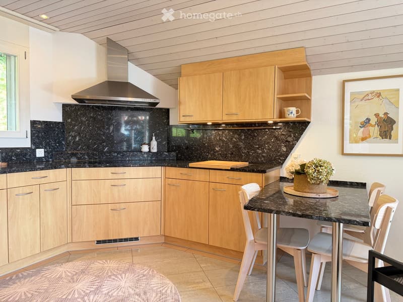 Modern kitchen with light wooden cabinets, dark granite countertops, a central island with seating, and a range hood above the stove.
