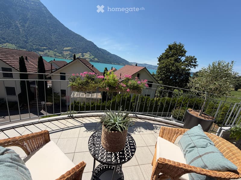 Balcony with wicker chairs, a small table, and potted plants, overlooking a scenic lake and mountain view.