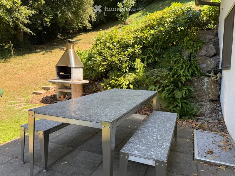 Outdoor patio area with a stone pizza oven, a grey stone table with metal legs, and matching benches, surrounded by lush greenery and a grassy lawn.