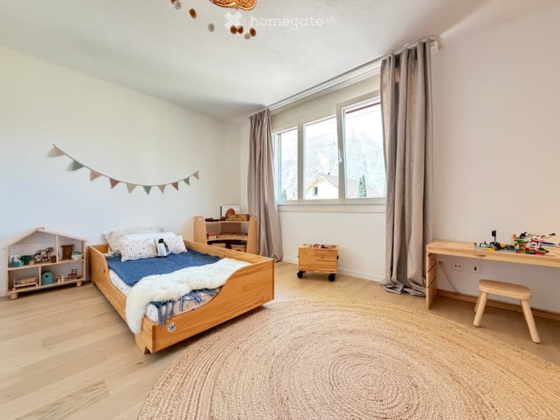 Cozy children's bedroom with a wooden bed, desk, and various toys, featuring large windows with curtains and a view of the mountains.