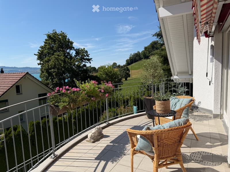 Balcony with wicker chairs, small table, potted plants, and scenic view of greenery and hills.
