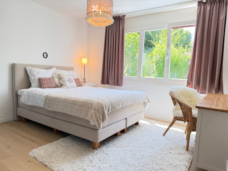 Cozy bedroom with a large bed, nightstand, table lamp, wicker chair with a fur throw, and large windows with curtains. The room features a white shag rug and wooden flooring.