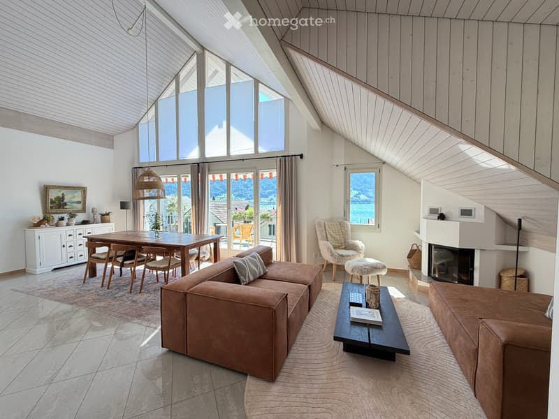 Bright living room with large windows offering a scenic view, featuring a brown sectional sofa, coffee table, dining area with a wooden table and chairs, a fireplace, and a cozy armchair.