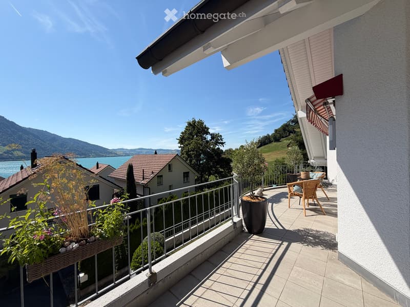 Balcony with a view of a lake and mountains, featuring a metal railing, potted plants, a wicker chair, and a small table.
