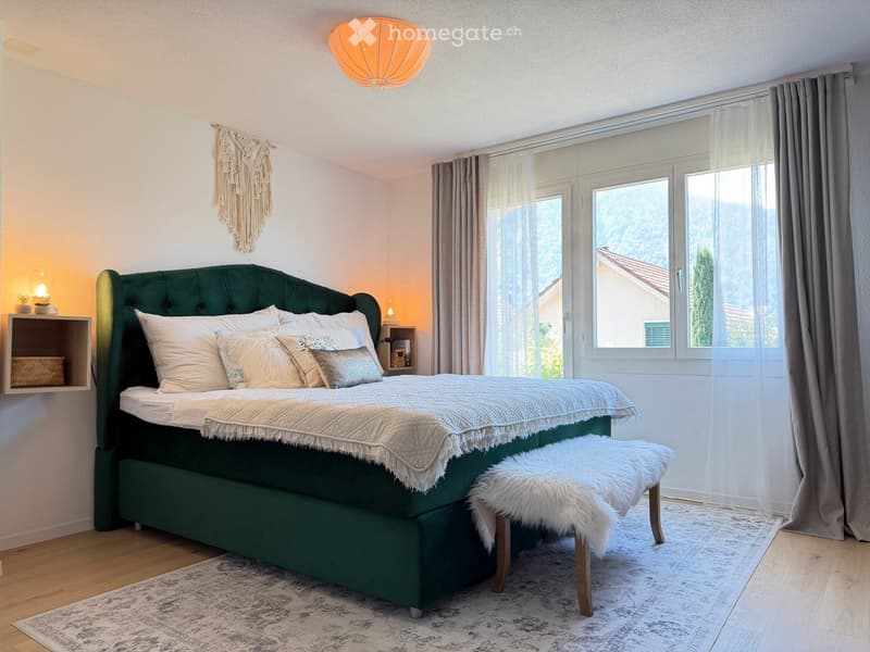 A well-lit bedroom with a green upholstered bed, white bedding, a fur-covered stool, and a nightstand with a lamp. Large windows with sheer curtains offer a scenic view.
