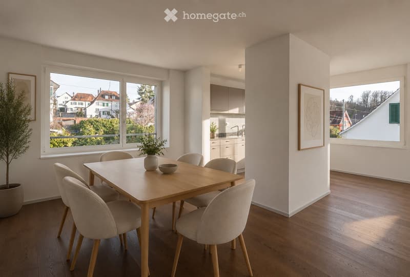 Modern dining area with a wooden dining table, six chairs, a vase with a plant, and a bowl on the table. The room features large windows with a view of the neighborhood, light-colored walls, and a small kitchen area visible in the background.
