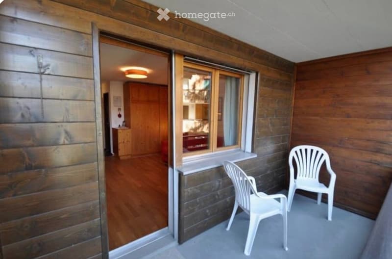 Balcony with wooden walls and flooring, two white plastic chairs, and an open doorway leading to an interior space with wooden cabinets and a visible light fixture.