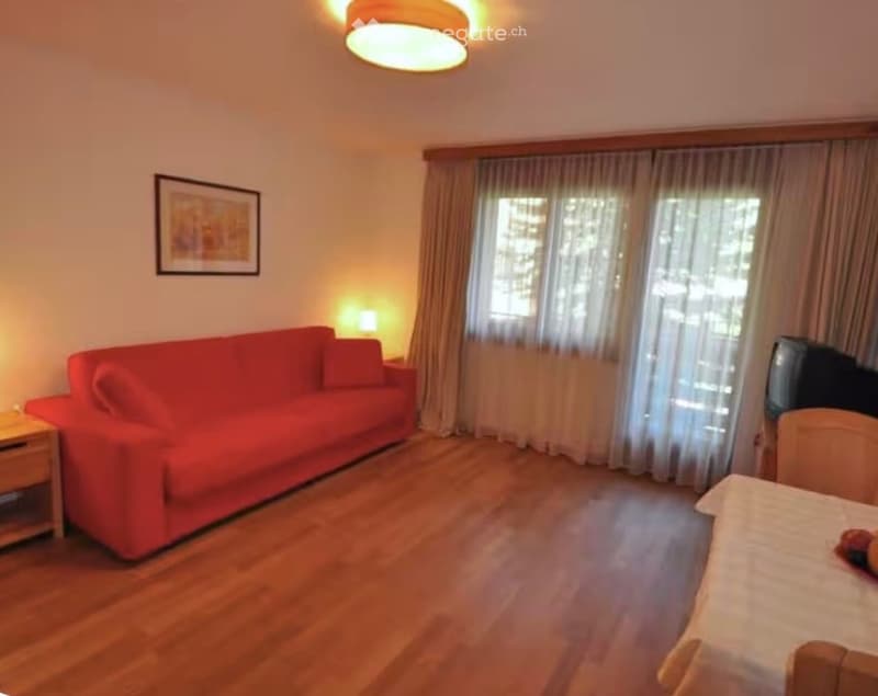 A cozy living room with a red sofa, wooden floor, a small table with a lamp, a television, and a picture frame on the wall. Large windows with curtains let in natural light.