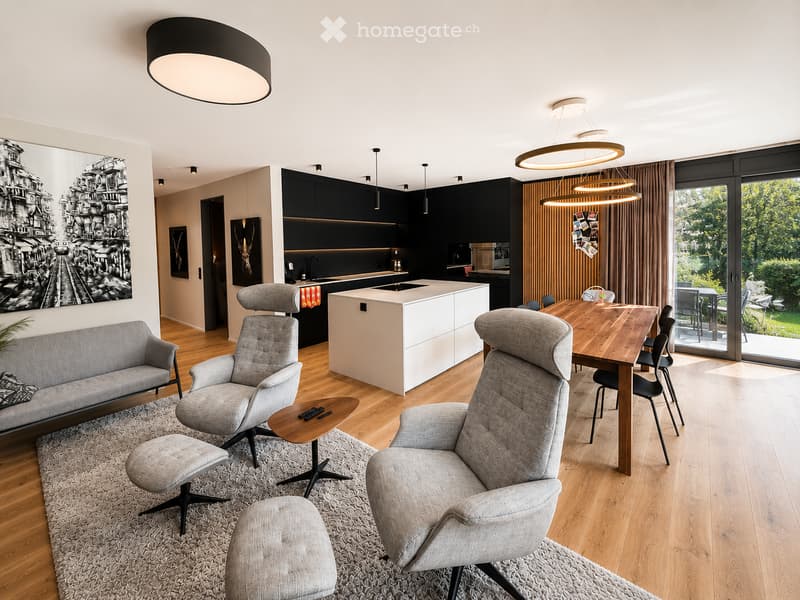 Open-plan living and dining area with modern furniture, including a sofa, chairs, a coffee table, a dining table, and a kitchen island. The space features wooden flooring, pendant lights, and large windows with curtains.