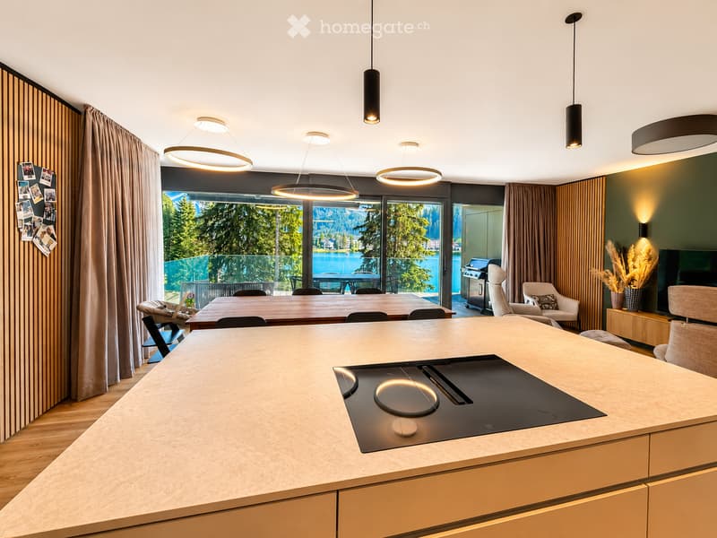 Modern kitchen with a large island featuring an induction cooktop, pendant lights, and a dining area with a table and chairs. A living area with a sofa and ottoman is visible, along with a scenic view of a lake and mountains through large windows.