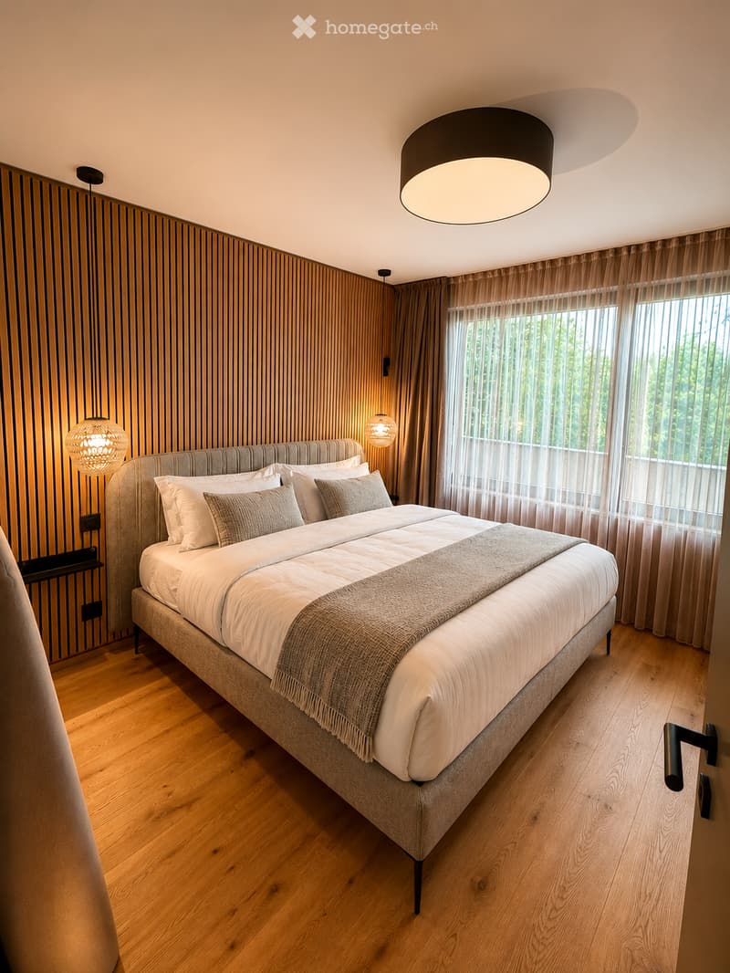 Modern bedroom with a large bed, multiple pillows, a blanket, wooden flooring, and large windows with curtains. The room features contemporary lighting fixtures and a sleek design.