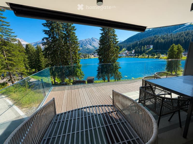 Balcony with wooden decking, glass railing, and outdoor furniture overlooking a lake and mountain landscape.