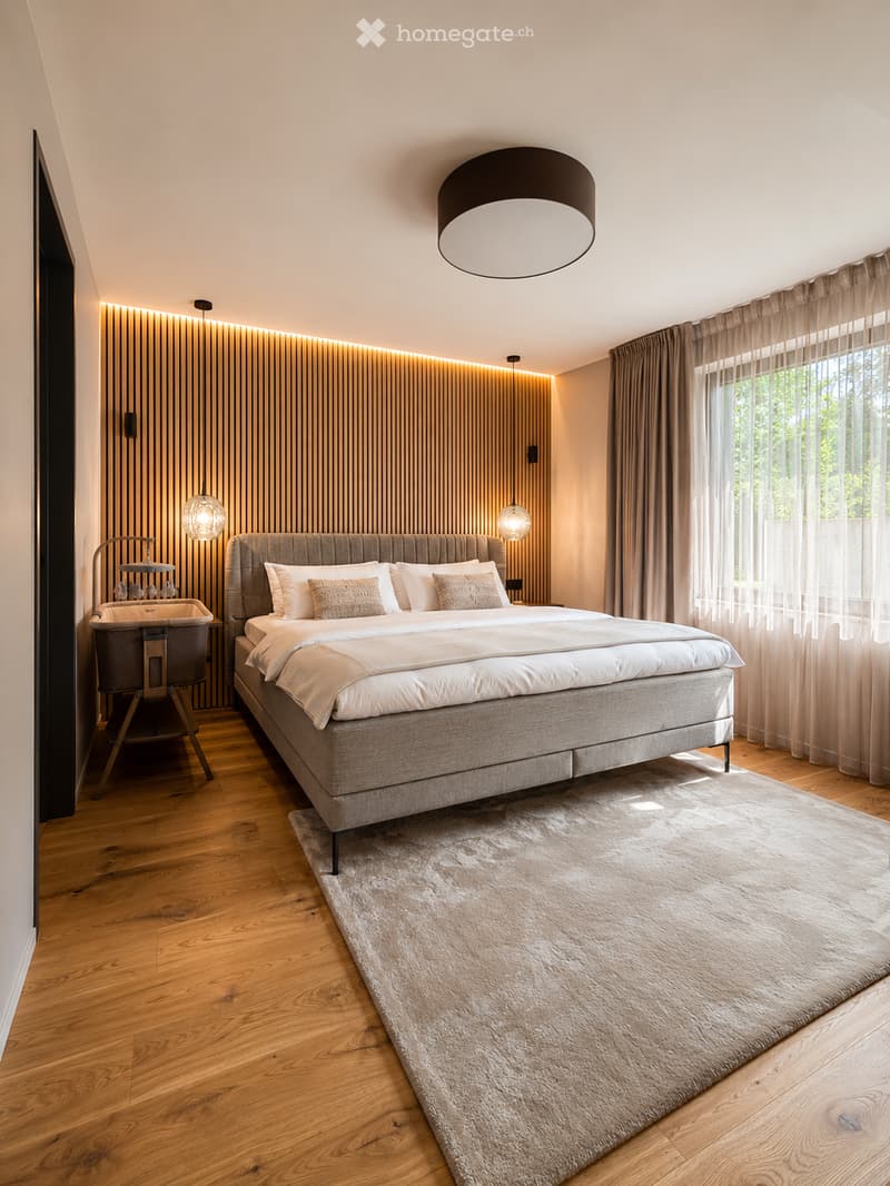 Modern bedroom with a large bed, nightstand, baby chair, and a round pendant light. The room features wooden flooring, a large window with curtains, and a textured rug.
