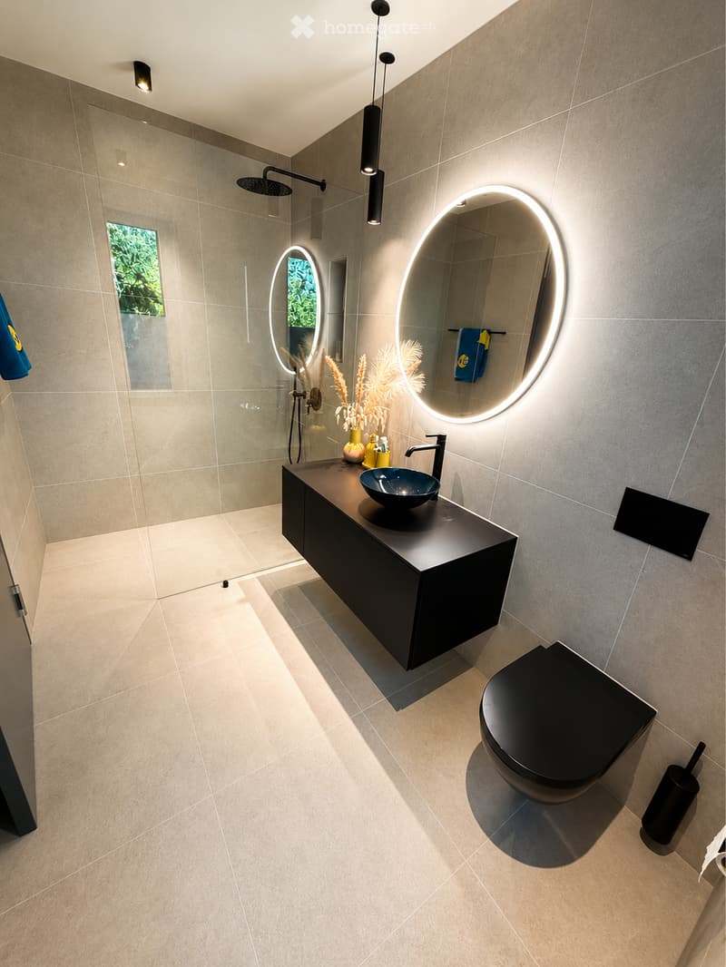 Modern bathroom with beige tiles, black vanity, sink, and toilet. Features a large illuminated mirror, a shower area, and decorative vase on the counter.