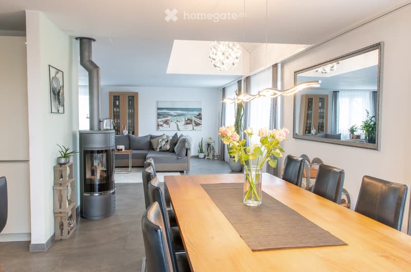 Open-plan living and dining area with a grey sectional sofa, wooden dining table with black chairs, modern fireplace, large mirror, and decorative vases with flowers.