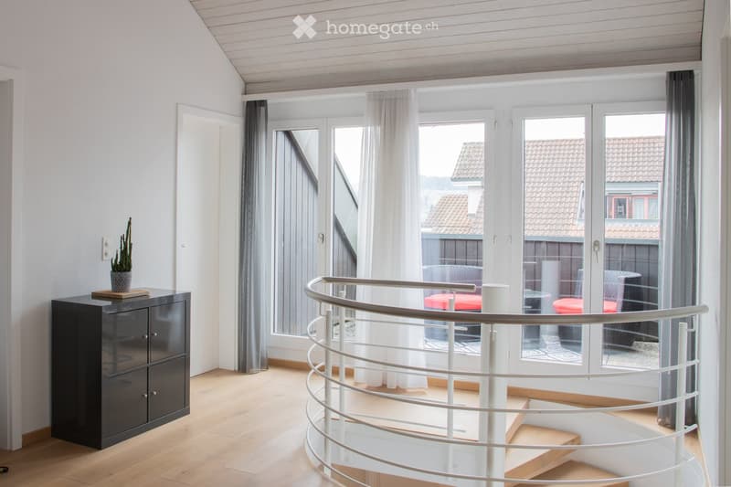 Spacious, modern room with light wooden flooring, white walls, and large windows with sheer curtains. A sleek black cabinet with a potted plant sits near the wall, and a curved white staircase with a glass railing leads upwards.