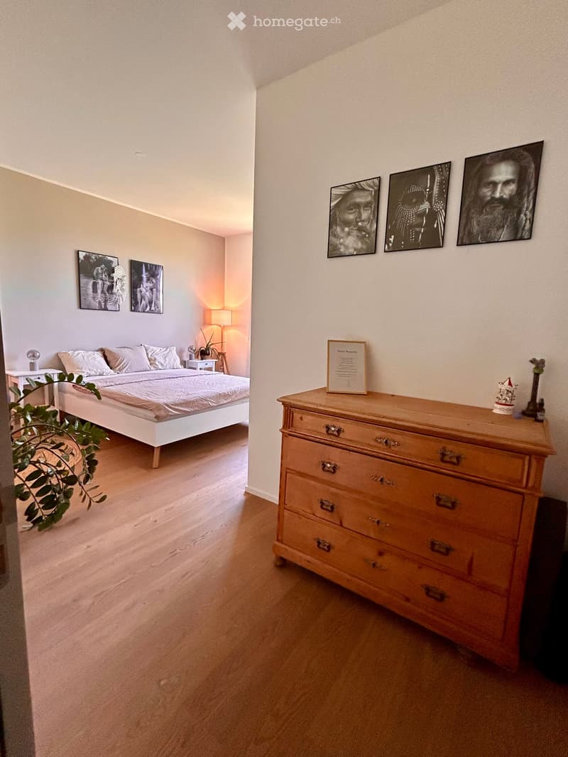 Bright bedroom with a white bed, wooden dresser, nightstand, lamp, and several picture frames on the walls.