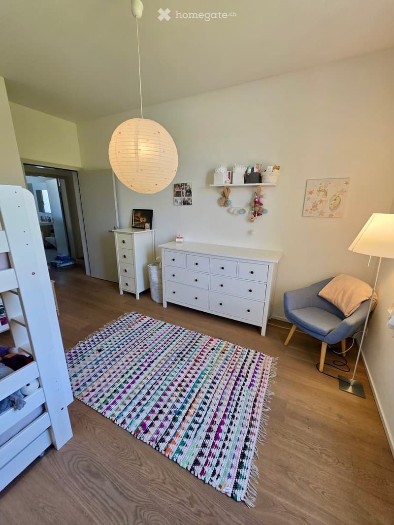 A well-lit room with wooden flooring, a colorful woven rug, a grey armchair, a white dresser, a hanging paper lantern, and shelves with decorative items.
