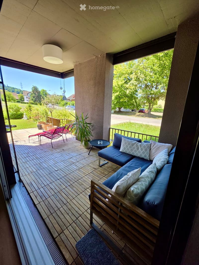 Balcony with wooden decking, blue sofa with patterned cushions, small round table, red chairs, and potted plants.