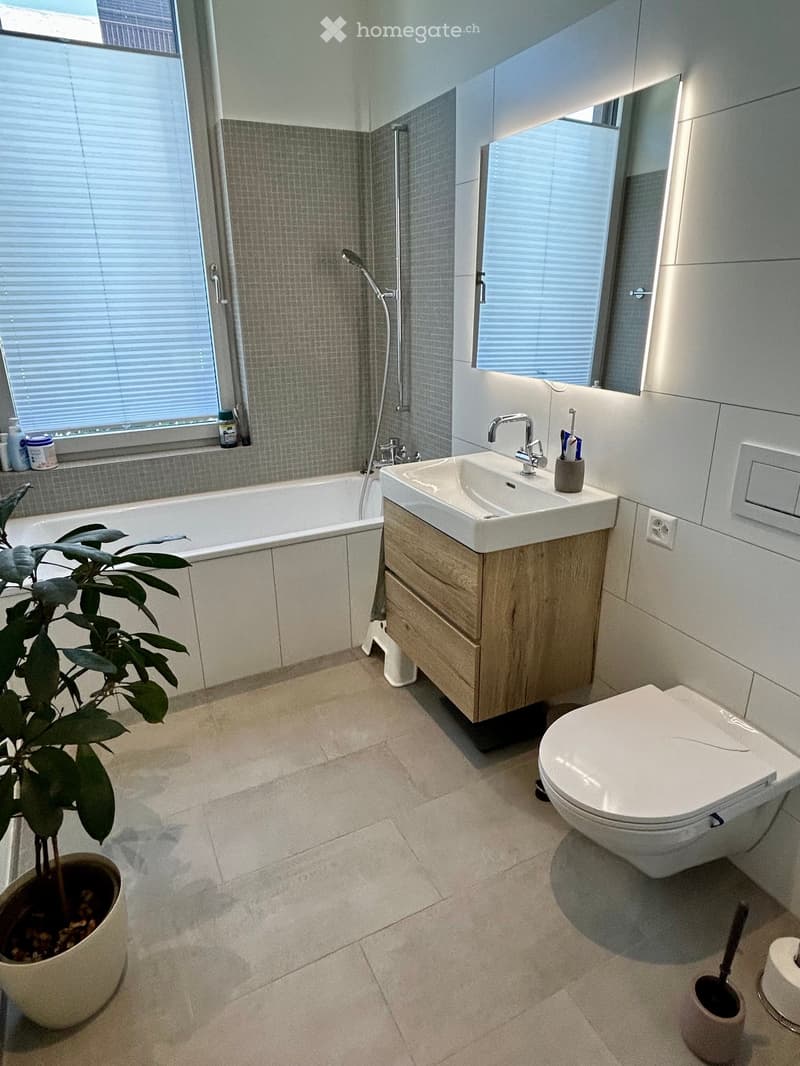 Modern bathroom with a bathtub, sink, toilet, and a small plant in a pot. The room features light-colored tiles and a large window with blinds.