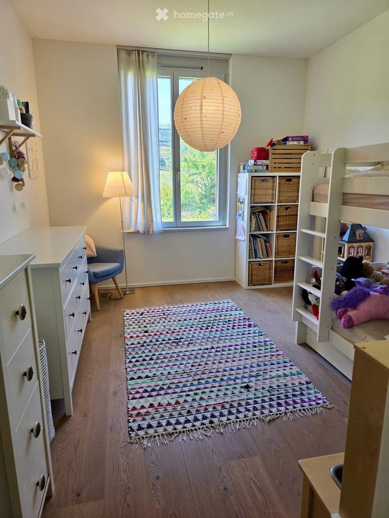 A well-lit bedroom with wooden flooring, a bunk bed, a colorful rug, a small table with a lamp, a shelf with storage boxes and books, and a chair. A window with curtains offers a view of greenery outside.