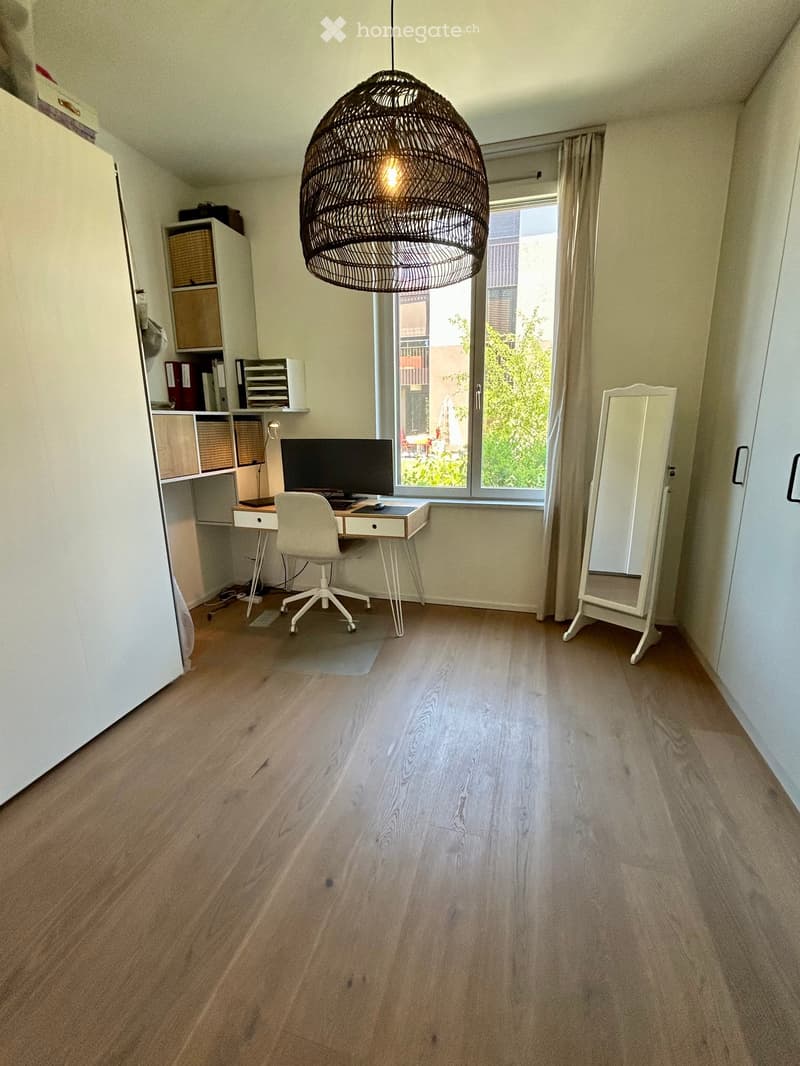 A modern, well-lit room with light wooden flooring, a large window with a view, a white desk with a computer monitor, a white office chair, a large round pendant light, a white mirror, and a storage unit with baskets.