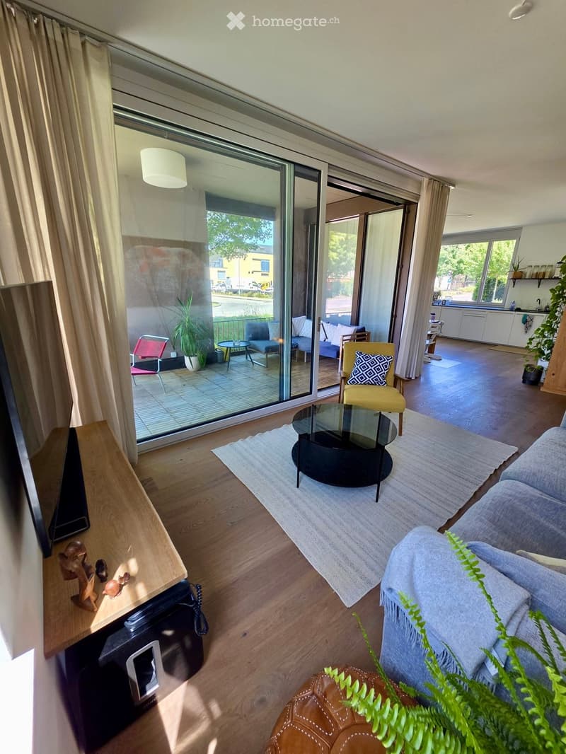 Modern living room with wooden flooring, large sliding glass doors leading to a balcony, sofa, coffee table, chairs, and plants.