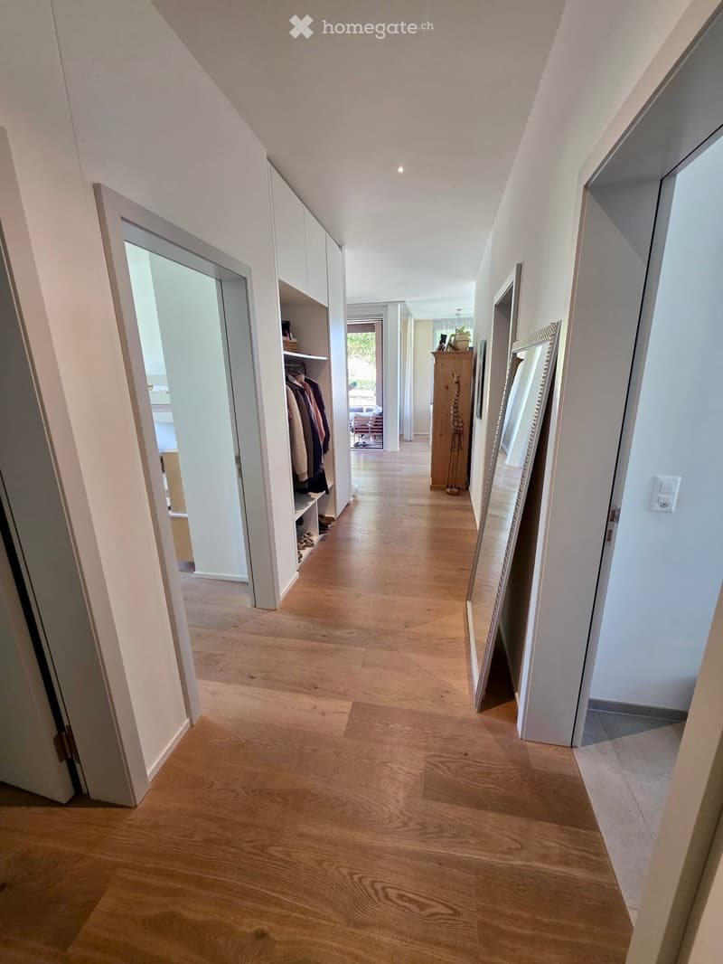 Bright hallway with wooden flooring, white walls, and built-in closet with hanging clothes. A large mirror leans against the wall, and a small wooden cabinet is visible in the background.