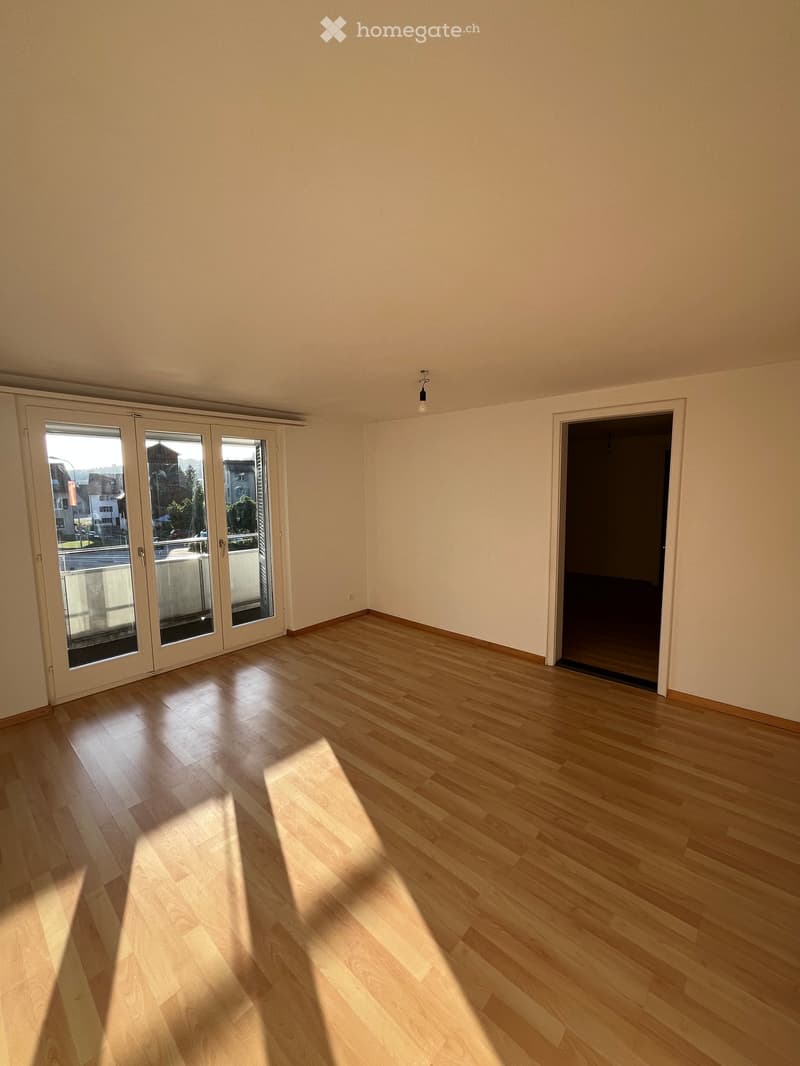 Empty room with light wooden flooring and white walls, featuring a large sliding glass door and a doorway leading to another space.