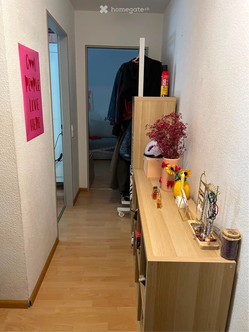 A narrow hallway with wooden flooring, a coat rack with jackets, a small table with decorative items, and a partially visible bedroom in the background.