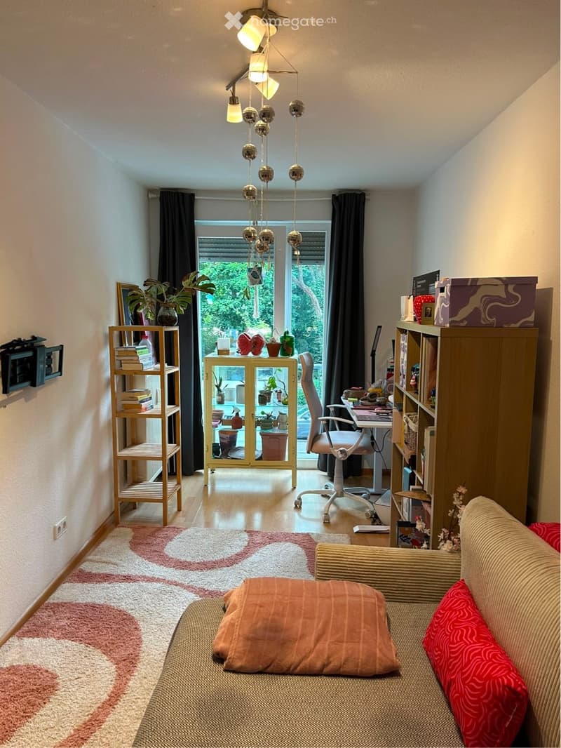 A cozy living room with a sofa, chair, small table, bookshelf, and decorative plants. The room features a colorful rug, modern lighting, and large windows with a view of greenery outside.