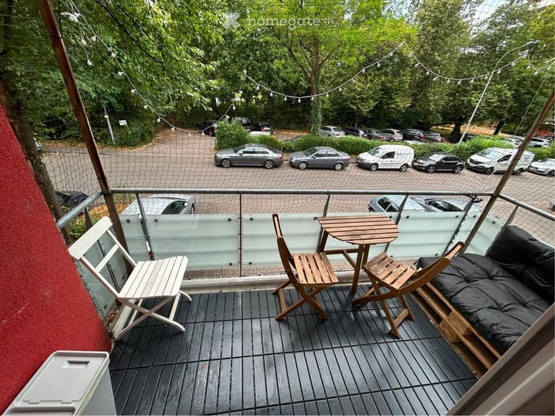 Balcony with wooden decking, glass railing, small table, chairs, and a sofa, overlooking a parking lot with cars and trees.