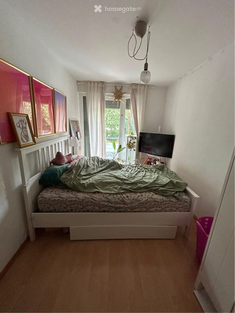 Cozy bedroom with a white wooden bed, green blanket, pillows, a television on a stand, a lamp, and framed pictures on the wall. The room has light-colored walls, wooden flooring, and a window with curtains.