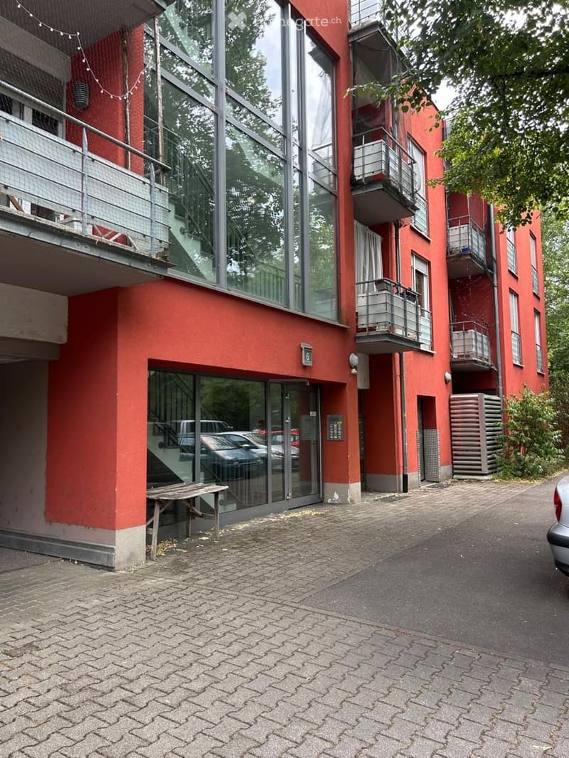 Red brick building with large glass windows and balconies, featuring a paved entrance area with parked cars.