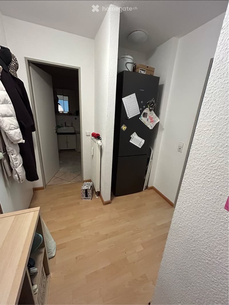 A narrow hallway with wooden flooring, white walls, and a coat rack on the left side. A refrigerator is visible on the right, with a doorway leading to a kitchen area in the background.