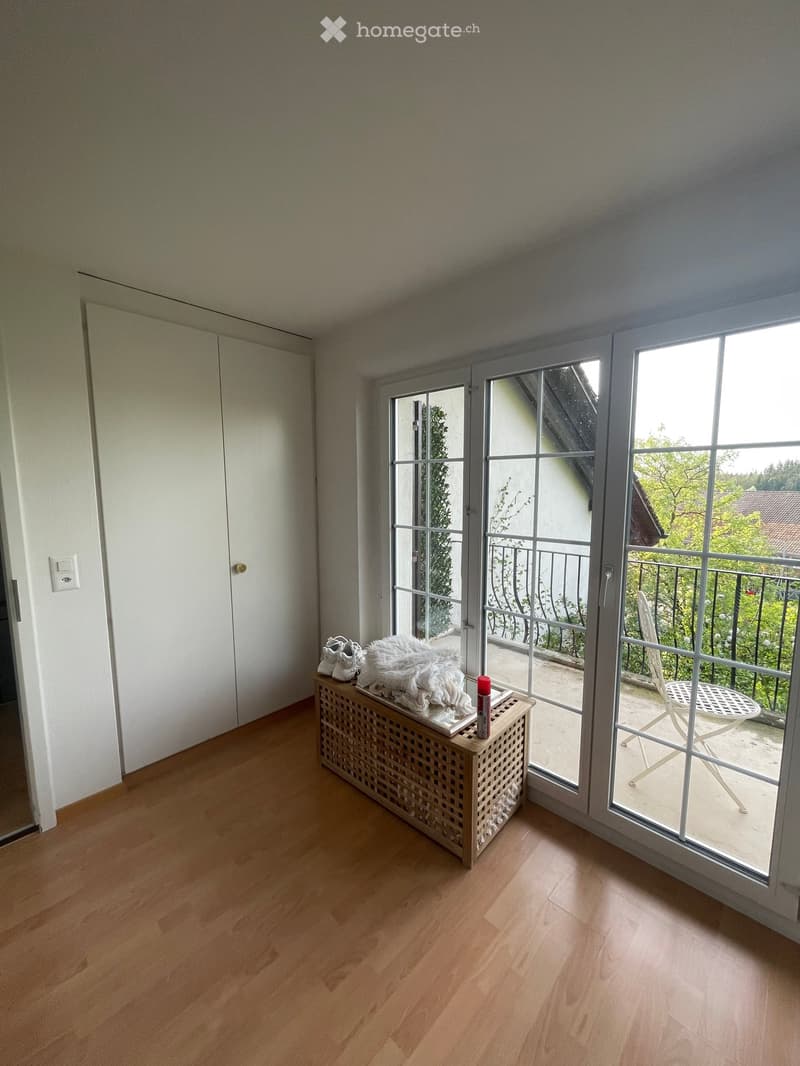 A small, well-lit room with wooden flooring, a bench with shoes and a fur throw, and a sliding glass door leading to a balcony with a chair and table.
