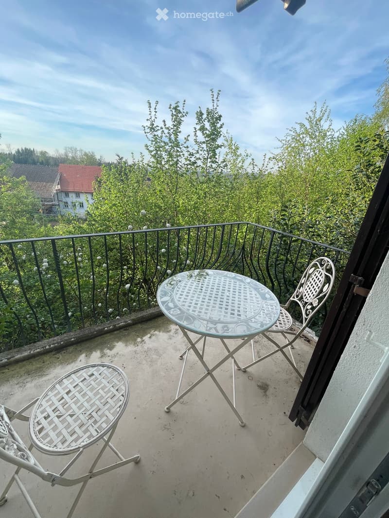 Balcony with a glass table and two white metal chairs, overlooking a green, leafy area with a house in the background.