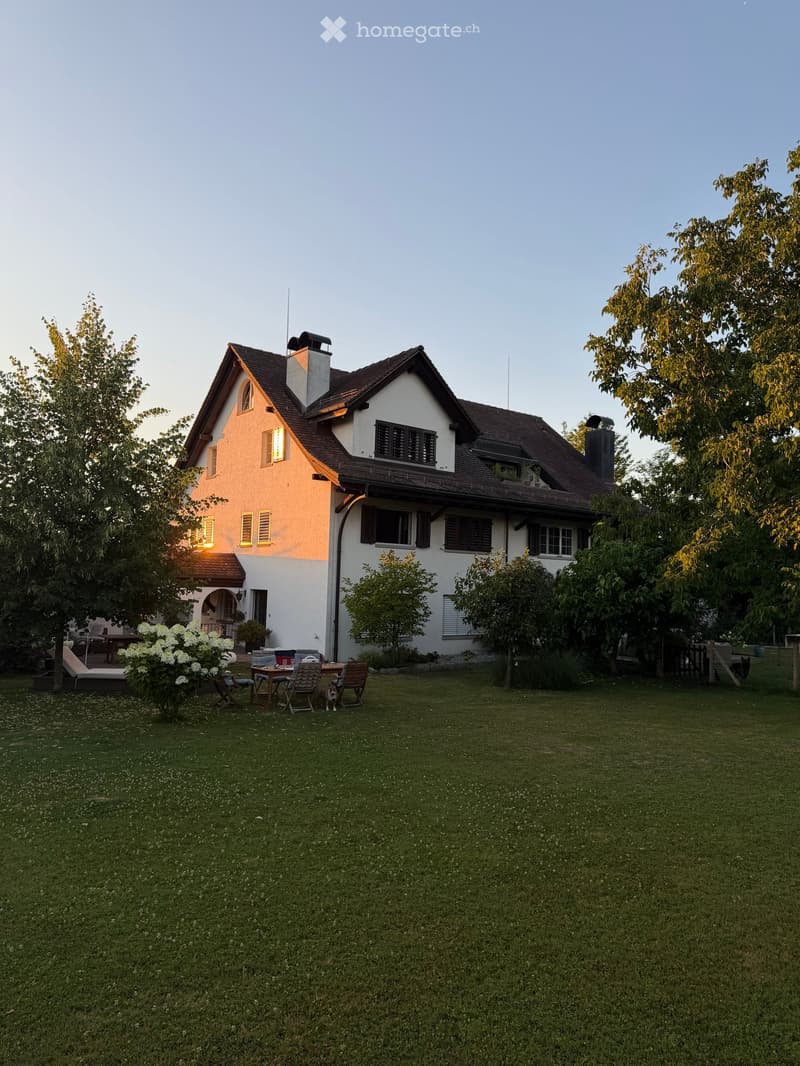 Two-story house with a well-maintained lawn, outdoor seating area, and garden. The house features a mix of white and beige exterior walls, multiple windows, and a chimney. The surrounding area includes trees and shrubs.