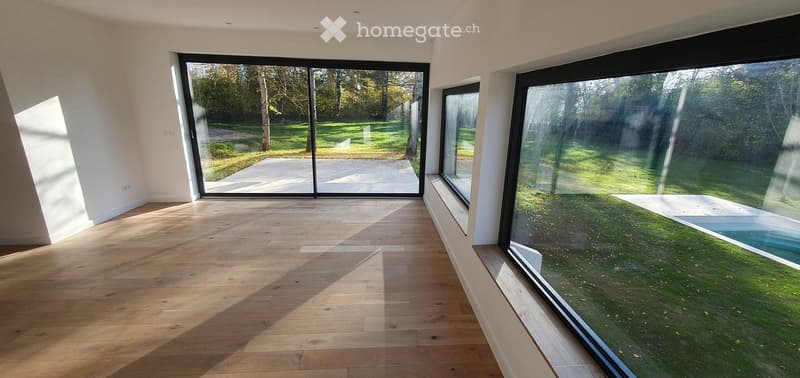 Empty room with wooden flooring and large windows overlooking a garden and pool area.