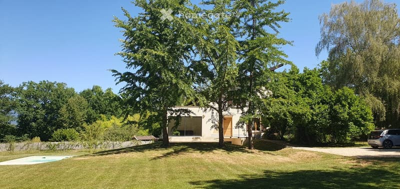 Modern two-story house with a swimming pool, surrounded by trees and greenery, with a parking area nearby.