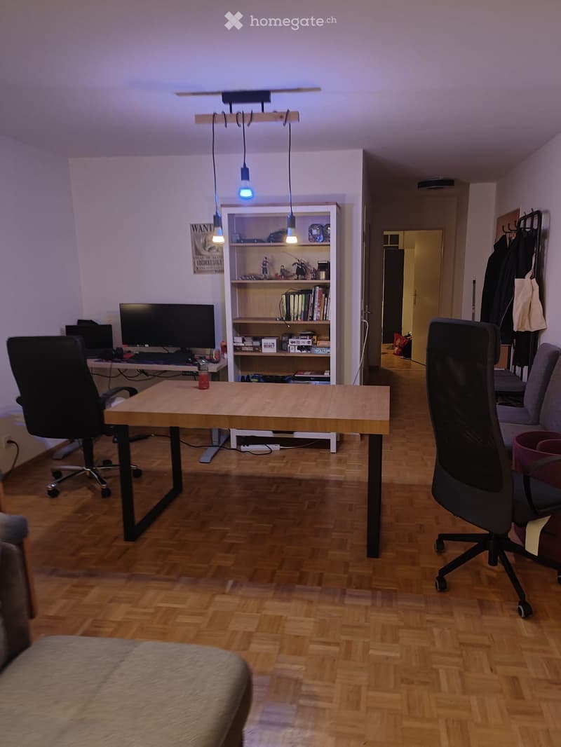 A modern, well-lit room with wooden flooring, a large wooden table, two office chairs, a sofa, a shelf with books and decorative items, and a computer setup with a screen and keyboard.