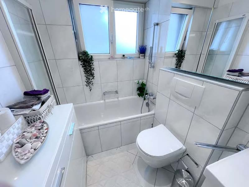 Modern bathroom with white tiles, bathtub, toilet, sink, mirror, and decorative elements including towels, a basket, and small plants.