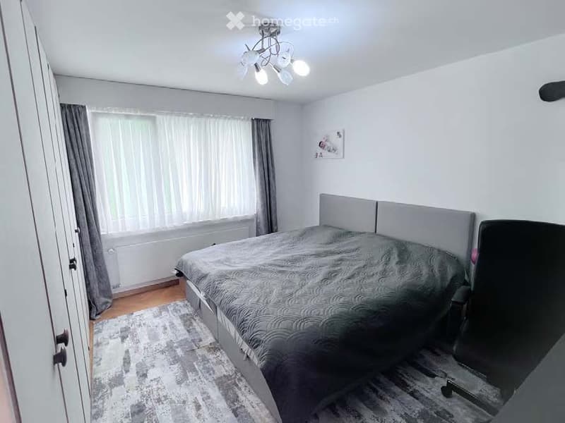 A modern bedroom with a large bed, headboard, nightstand, chair, picture frame, and a curtained window. The room features a patterned rug and a ceiling light fixture.