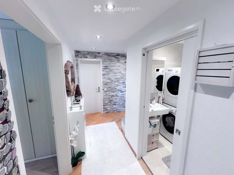 Hallway leading to a bathroom and laundry area, with a tiled accent wall, wooden flooring, and a small plant in the corner.
