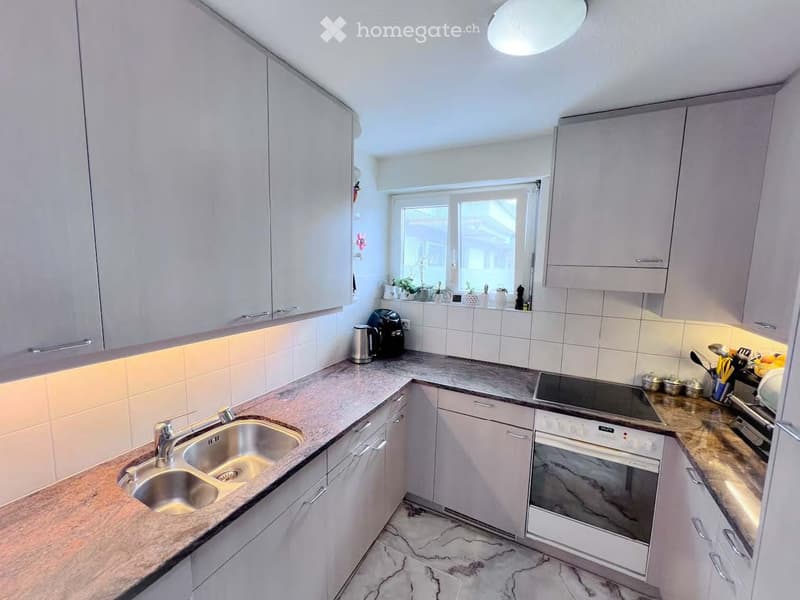 Modern kitchen with light grey cabinets, marble countertops, and stainless steel appliances including a sink, oven, and stove. The space is well-lit with natural light from a window and has a sleek, contemporary design.