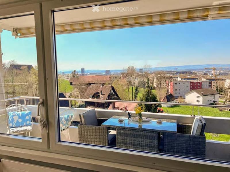 Balcony with outdoor furniture, overlooking a scenic cityscape with houses, greenery, and distant mountains.