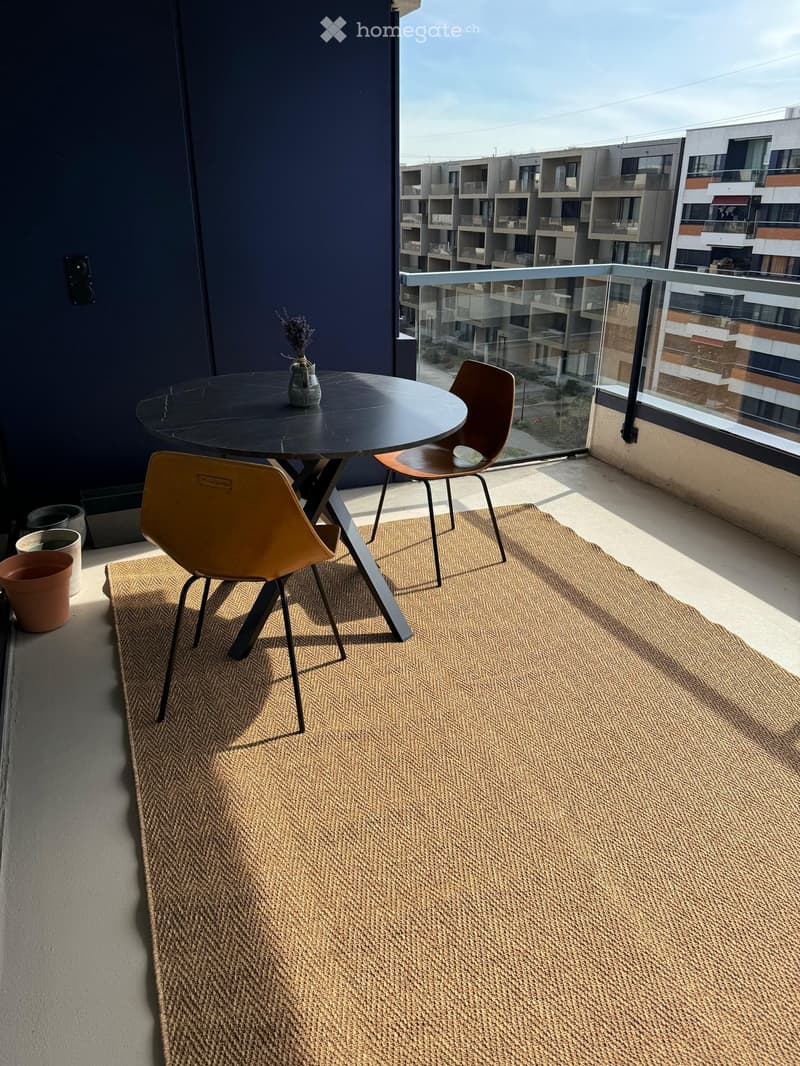 Balcony with a round black table, two modern chairs, a small vase with dried flowers, a large woven rug, and a glass railing overlooking a modern apartment complex.