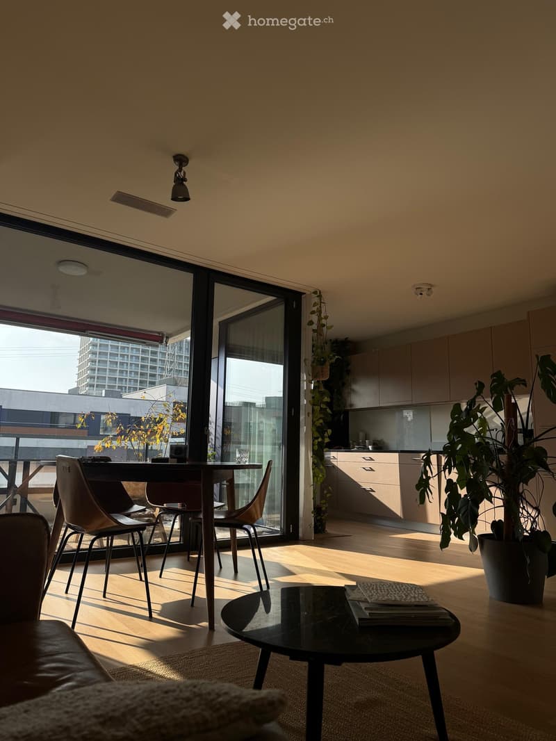 Bright living and dining area with a sofa, coffee table, dining table, chairs, and kitchen area visible through an open sliding glass door.
